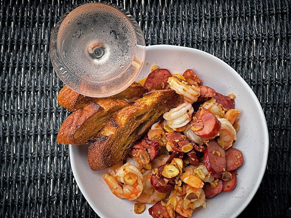 a white bowl of shrimp and chorizo beside a glass of chilled rose on a black wicker surface